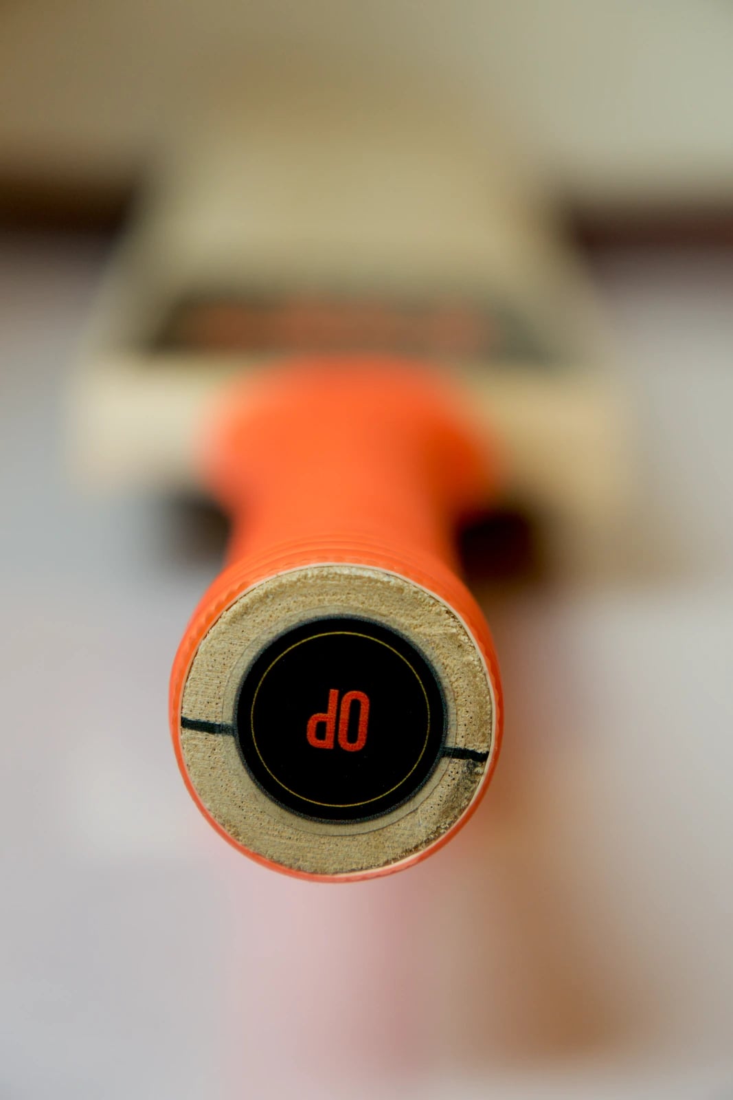 Close-up looking down the handle at its end, a black cap with a red OP logo ringed by the wood of the handle and the orange grip, with the blade out of focus behind